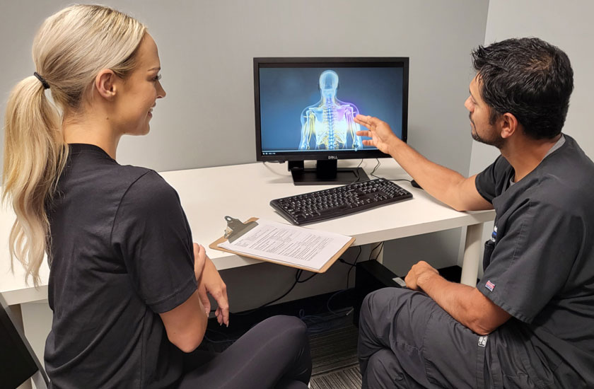 A Chiro One chiropractor points to a digital spinal scan on a monitor while explaining treatment to a patient.