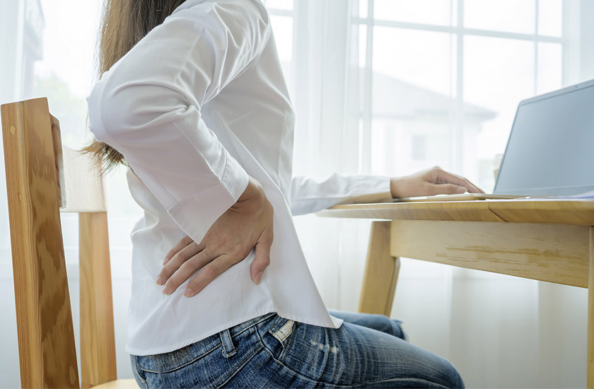 A woman sitting at a desk and holding her lower back in discomfort while working on a laptop, illustrating common symptoms of lower back pain from poor posture or long hours of sitting.