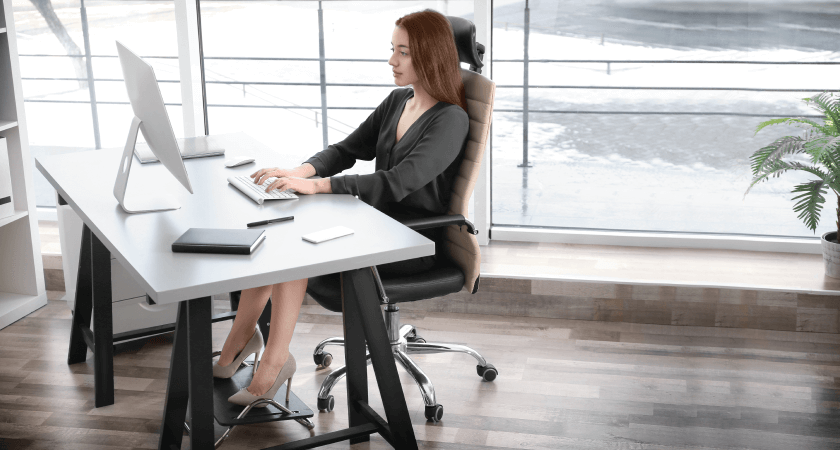 A woman working at her desk with proper ergonomics—seated in an ergonomic chair, using a footrest, and viewing a monitor positioned at eye level