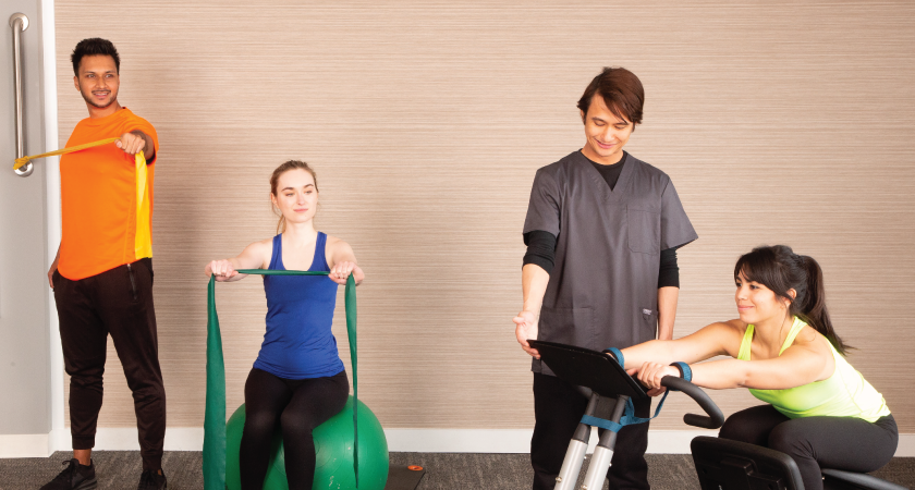 A chiropractic technician assists three individuals during a therapy session, with one person using a resistance band, another on an exercise ball with a resistance band, and a third on a Precor stretching machine.