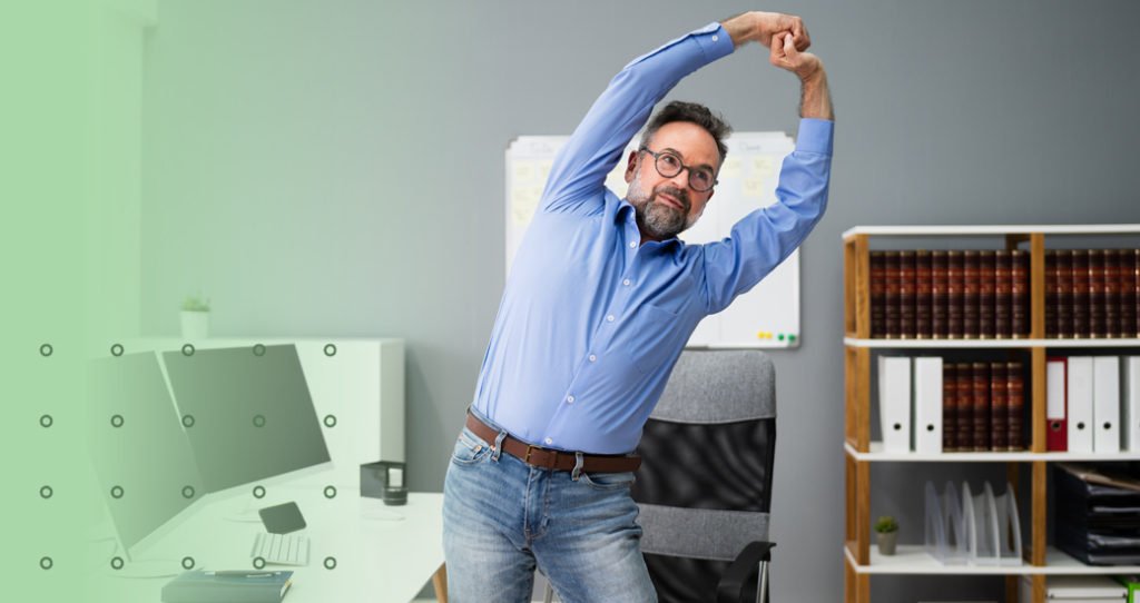 A middle-aged man in a blue dress shirt and jeans performs a side-bending stretch with arms overhead in a professional office, illustrating workplace exercises or daily habits to reduce back pain.