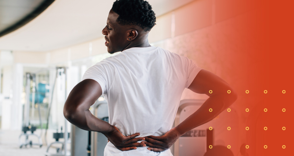 A muscular man in a white t-shirt stands in a gym, grimacing while holding his lower back with both hands, indicating severe low back pain or a disc problem.