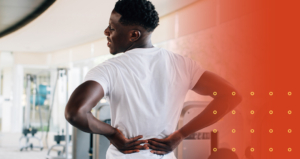 A muscular man in a white t-shirt stands in a gym, grimacing while holding his lower back with both hands, indicating severe low back pain or a disc problem.