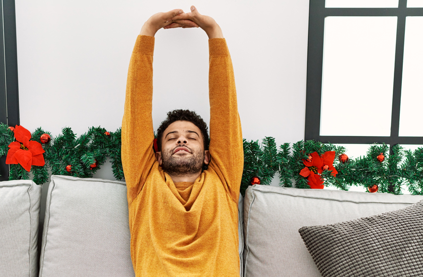 A relaxed man in an orange sweater stretches his arms over his head while sitting on a sofa, eyes closed, with a Christmas garland decorated with red poinsettias behind him, illustrating stress relief during the holidays.