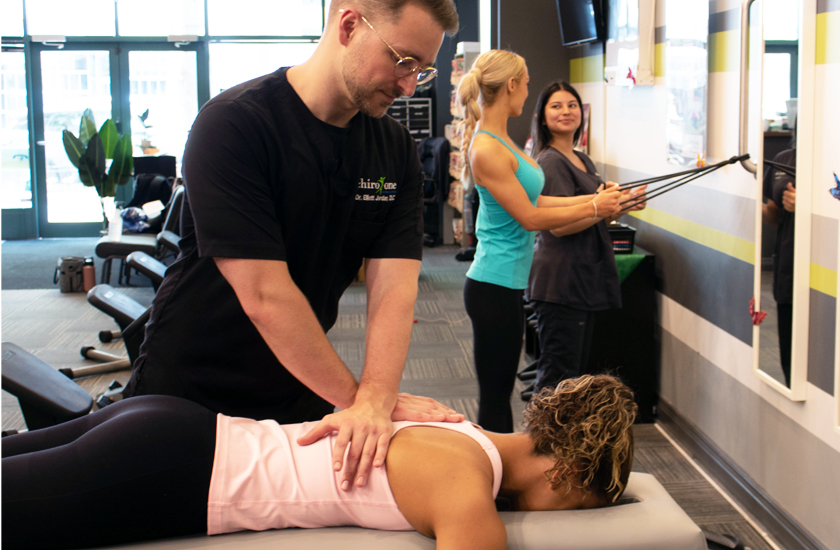 A male chiropractor in glasses performing a manual chiropractic adjustment on a female patient's middle back while she lies prone on a table, with another patient exercising in the background.
