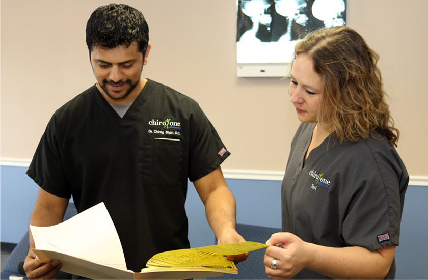 A male chiropractor and female clinic staff, wearing medical scrubs, are reviewing patient files and paperwork, with an X-ray light box visible in the background, representing integrated healthcare collaboration.