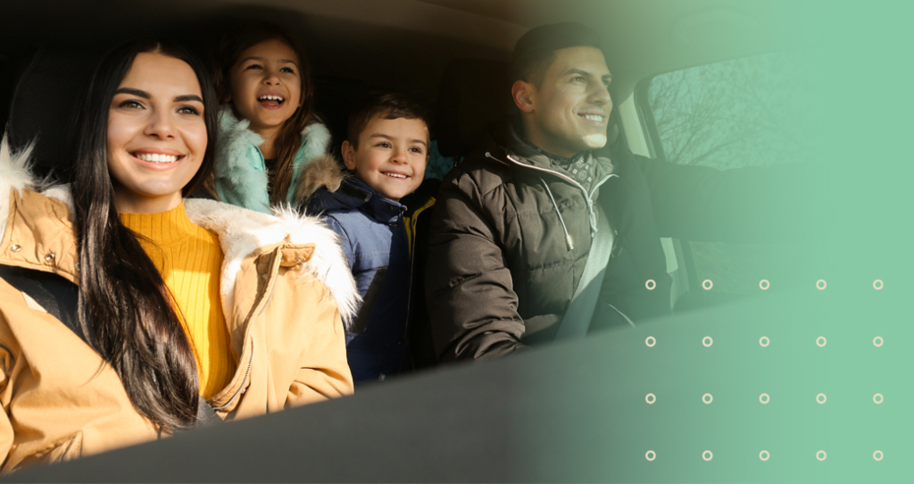 A happy family of four—mother, father, and two young children (a girl and a boy)—are seen smiling and laughing while riding together in a car. They are warmly dressed in winter coats, suggesting they are traveling for the holidays or on a winter road trip.