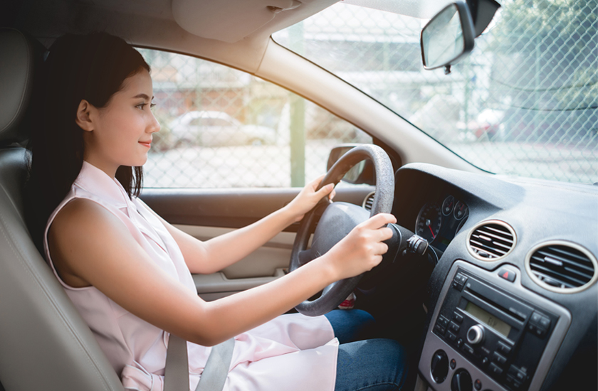 A young woman with long dark hair, wearing a light pink sleeveless top and jeans, is driving a car. She is looking straight ahead, with both hands on the steering wheel, suggesting she is focused on her journey or on a holiday road trip. The sun is shining through the side window.