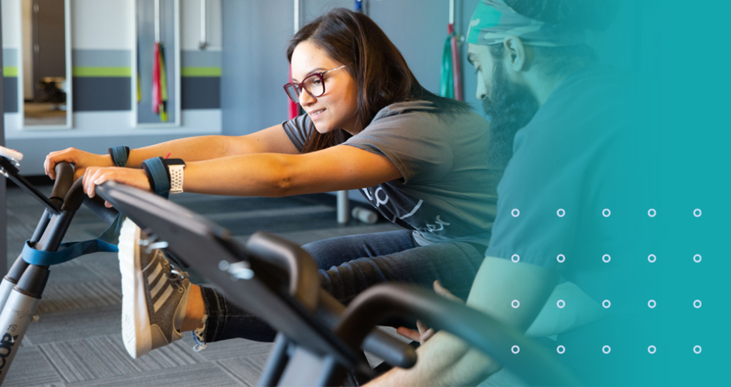 A woman smiling as she performs an activetherapy on rehabilitation equipment, guided by a male chiropractic technician, demonstrating active pain management and physical therapy.