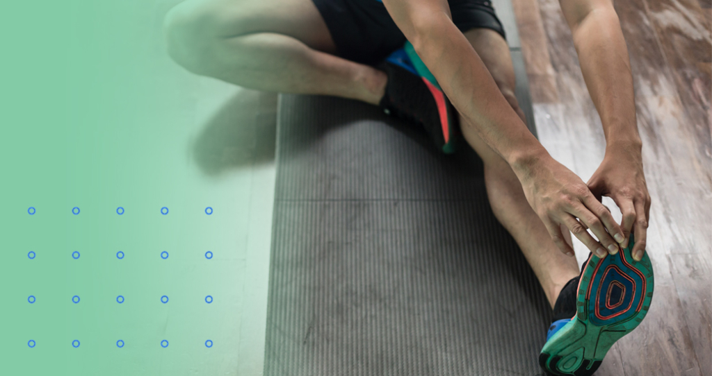 A close-up of a person sitting on a mat, performing a seated toe touch or hamstring stretch by reaching for their foot, illustrating flexibility or post-workout cool-down.