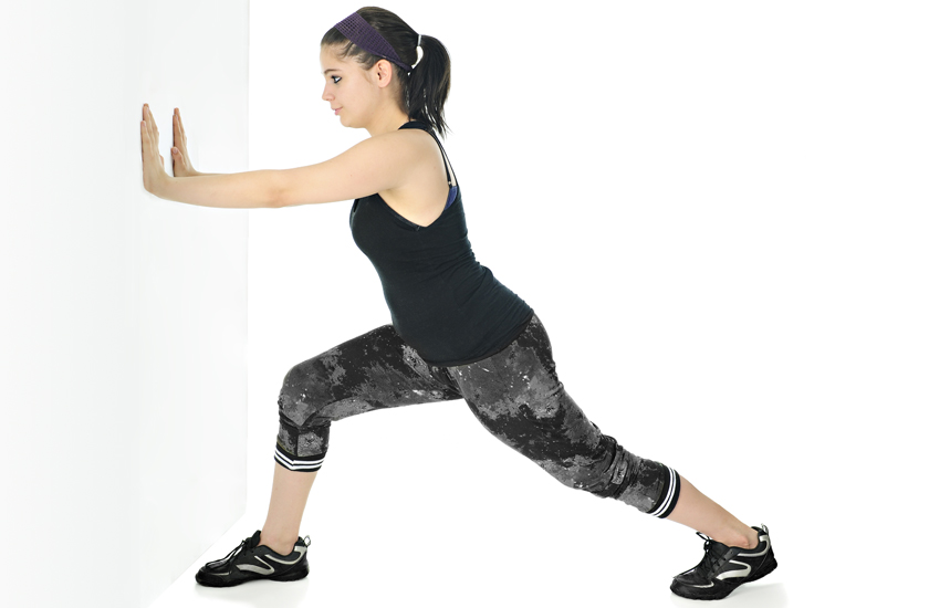 A young woman performs a standing calf stretch or gastrocnemius stretch against a white wall, with one leg extended back, demonstrating lower leg flexibility exercises.