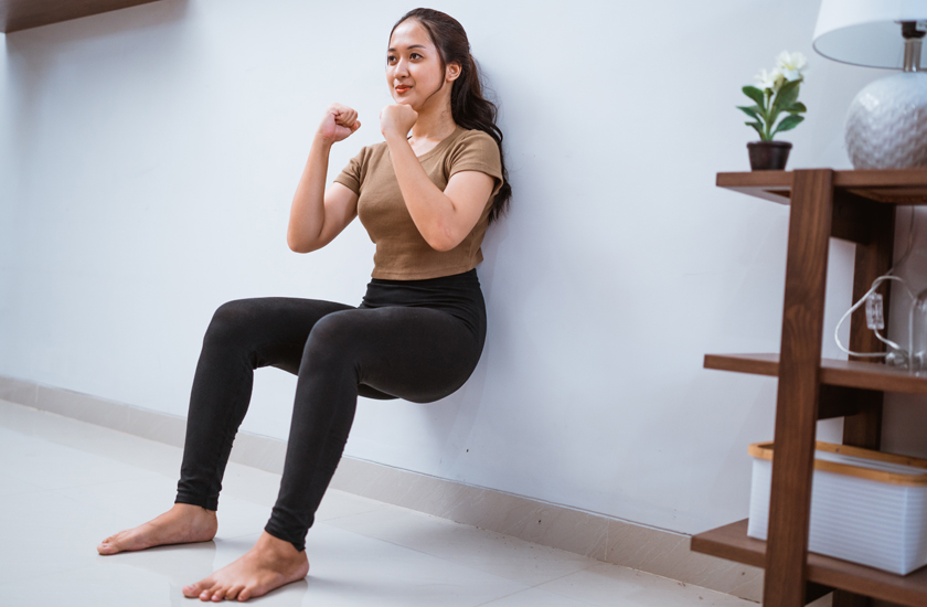 A young woman performs a wall squat or wall sit exercise, maintaining a 90-degree angle with her knees to build quadriceps and lower body strength.