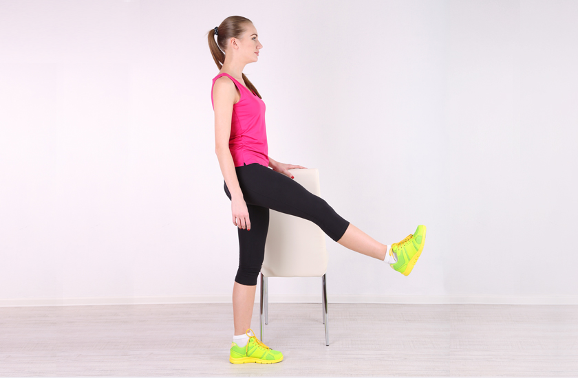 A woman stands next to a chair, performing a standing leg raise or quadriceps strengthening exercise by lifting one leg straight forward, demonstrating a balance and lower body workout.