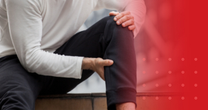 A man wearing a white long-sleeve shirt and black sweatpants sits and clutches his knee and upper hamstring/thigh area, illustrating knee pain, piriformis syndrome, or sciatica pain.