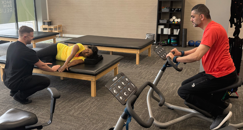 A chiropractor provides manual therapy to a patient, while another patient exercises on a rehabilitation machine in a Chiro One clinic.
