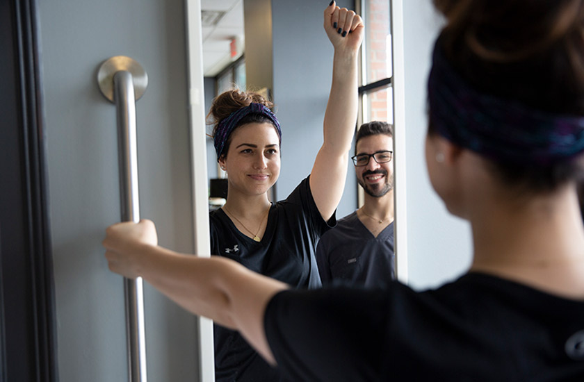 A Chiro One patient performing guided range-of-motion exercises in front of a mirror while a chiropractor observes her spinal alignment and arm mobility.